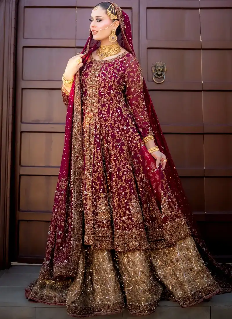 Burgundy front-slit bridal gown standing view with ornate gold borders and a bronze-gold lehenga