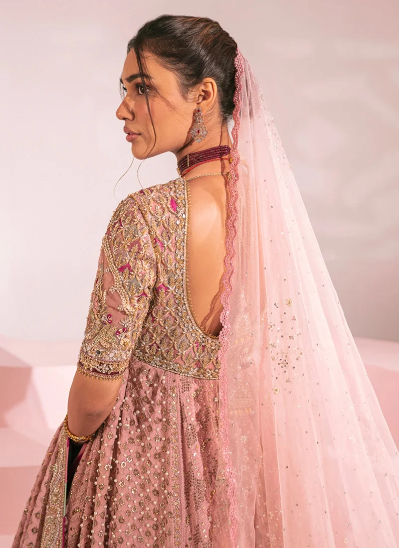 Dusty pink bridal pishwas in a seated front view, showing its wide embroidered paisley border.