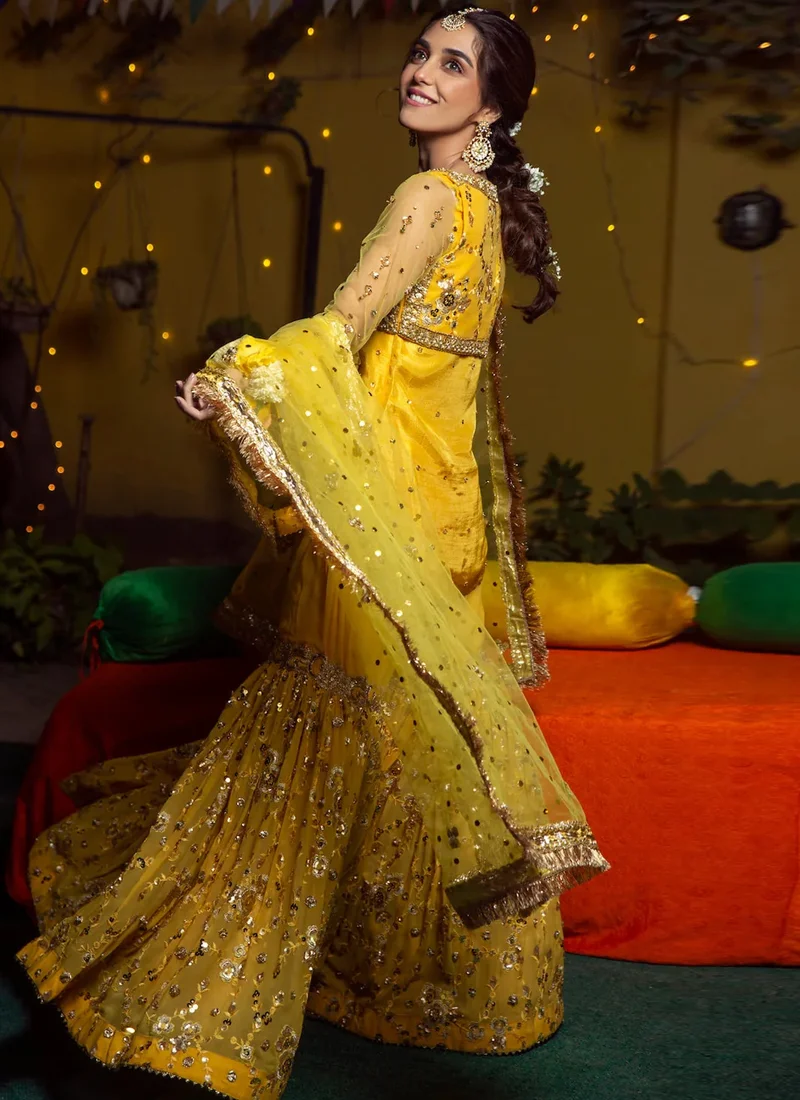 Yellow mehndi gharara suit seated front view with gold floral motifs and a fringed net dupatta
